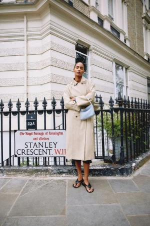 Model standing outdoors on a stone pavement wearing a beige trench coat and holding a light blue shoulder bag