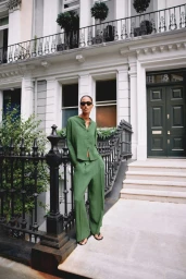 Model standing outdoors wearing a green button-up shirt and matching trousers in front of a white building with black railings.