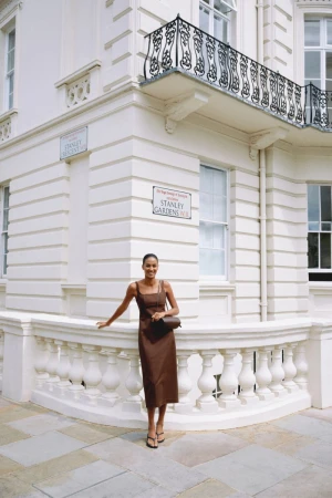 Model standing outdoors wearing a brown midi dress against a white residential building exterior with wrought-iron balcony.