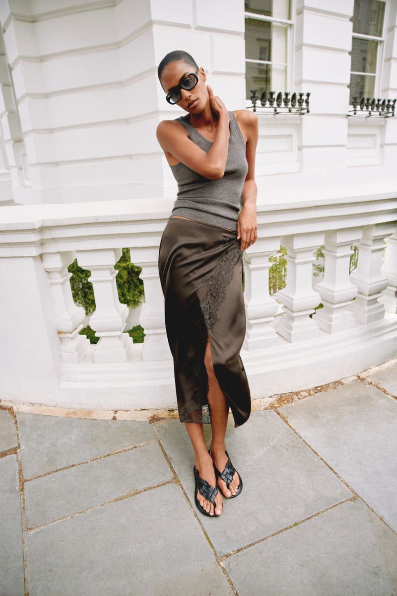 Model wearing a grey tank top and brown silk skirt leaning against a white classical stone balustrade