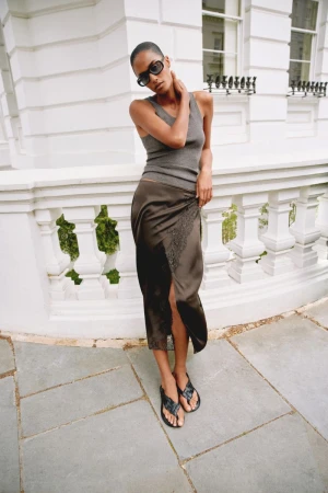 Model wearing a grey tank top and brown silk skirt leaning against a white classical stone balustrade