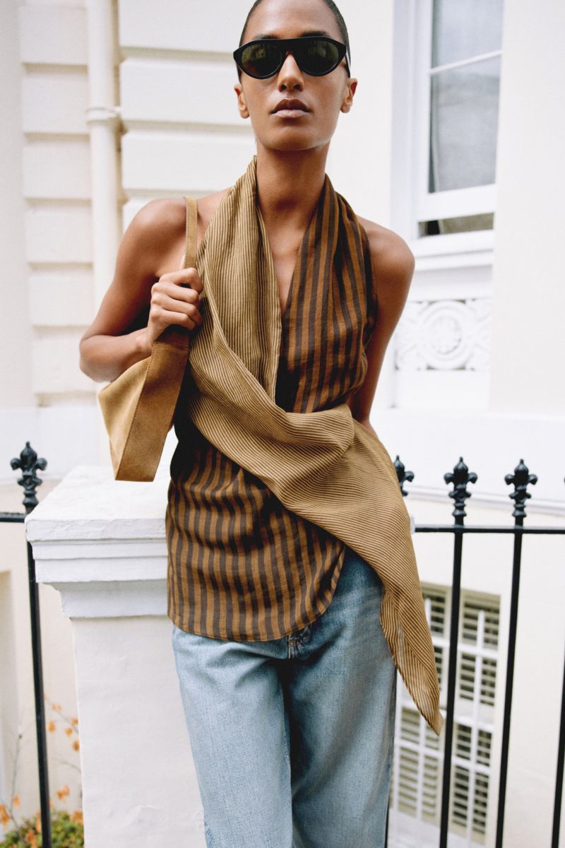 Model in sunglasses and striped top holding a shoulder bag in front of a white building exterior