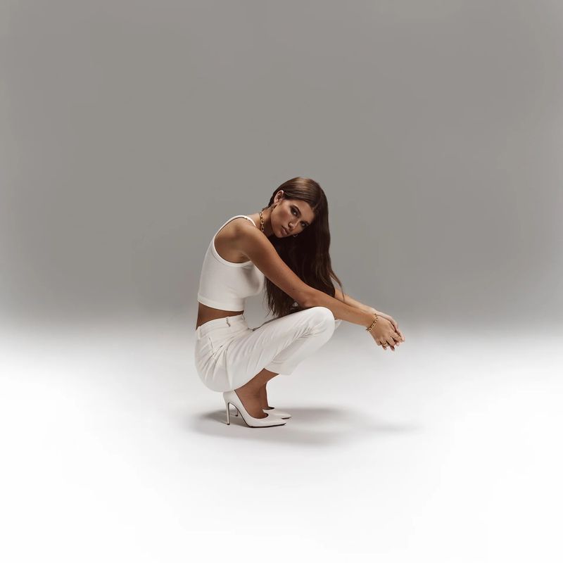 Brunette woman in a crouched pose, wearing a white crop top, white pants, and heels, against a gradient gray backdrop in a photo studio.