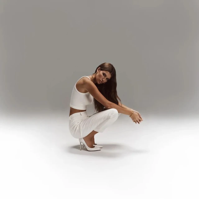 Brunette woman in a crouched pose, wearing a white crop top, white pants, and heels, against a gradient gray backdrop in a photo studio.