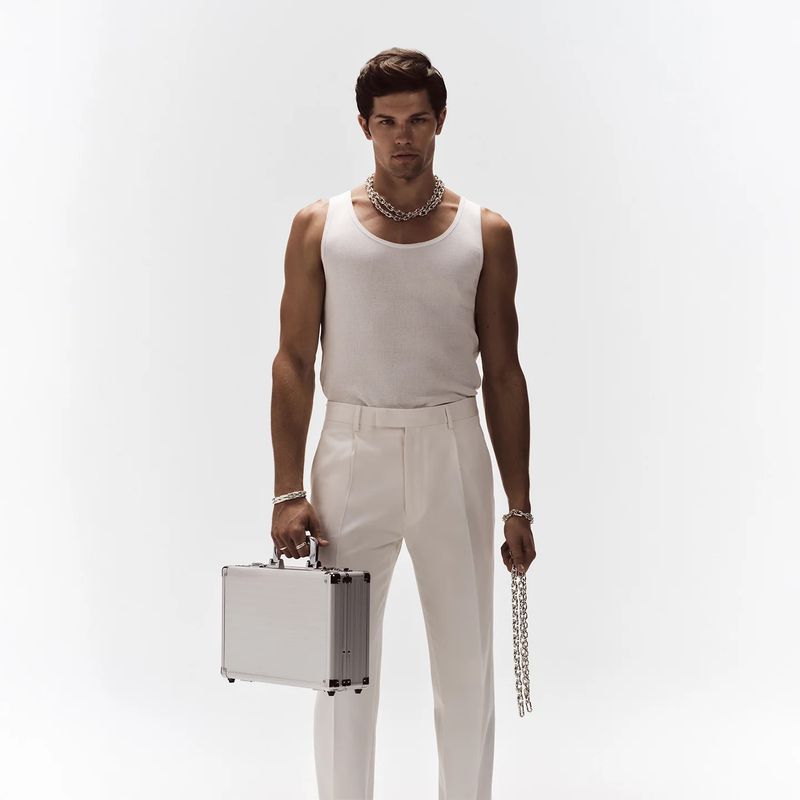 A man with short dark hair, wearing a white tank top and pants, poses with a metal briefcase and a silver chain against a white backdrop.