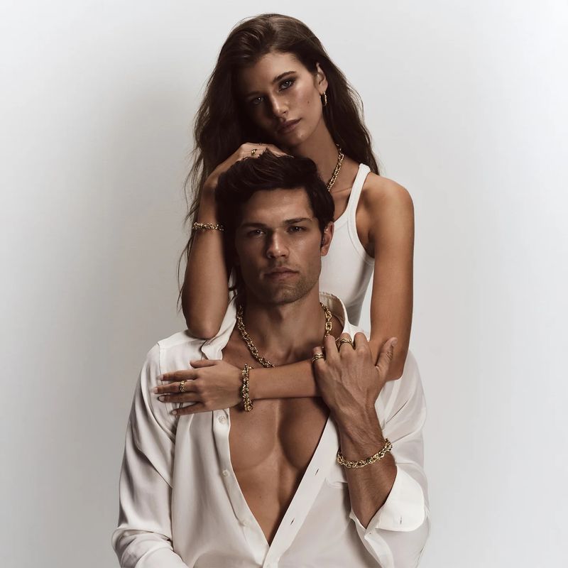 Attractive couple poses indoors for a jewelry advertisement. The man wears an open white dress shirt, and the woman wears a white tank top.