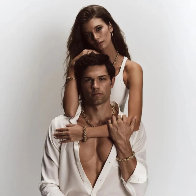 Attractive couple poses indoors for a jewelry advertisement. The man wears an open white dress shirt, and the woman wears a white tank top.