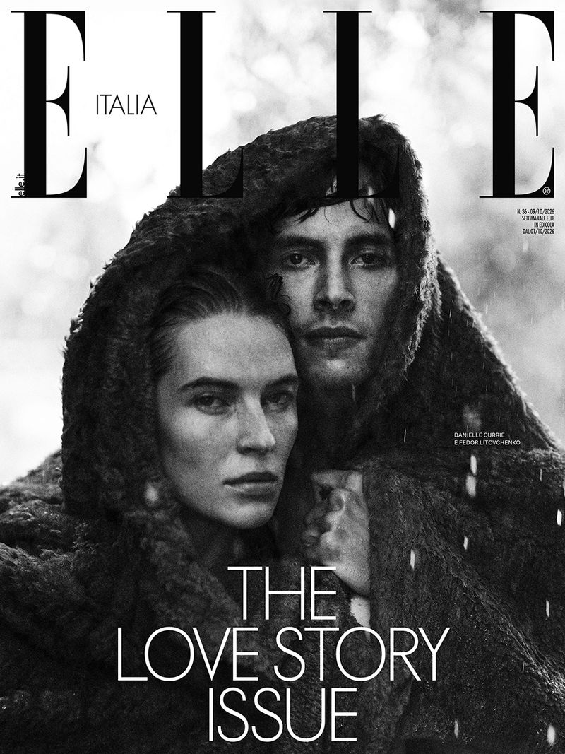 Monochrome magazine cover featuring a woman and man wrapped together in a heavy textured dark hooded garment outdoors.