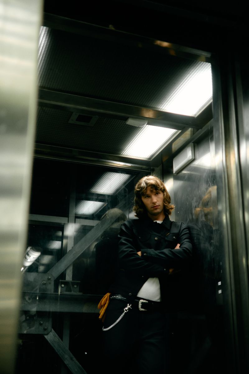 Model standing inside an industrial elevator wearing a dark denim jacket and white t-shirt for Wrangler X Slam Jam.