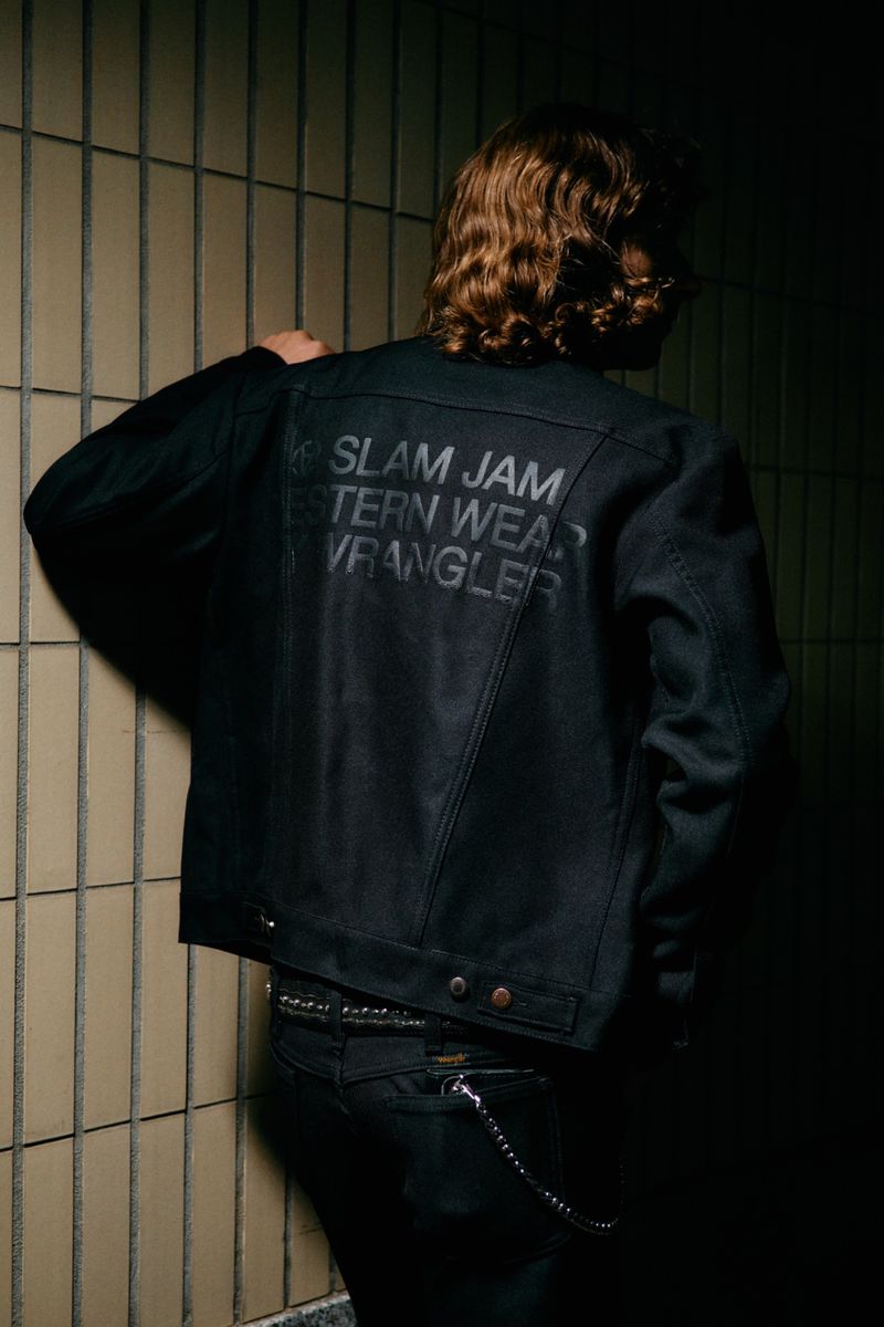 Model wearing a black denim jacket with Slam Jam and Wrangler lettering on the back against a tiled wall