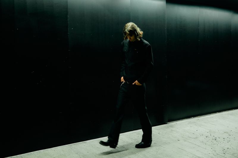 Model walking alongside a dark wall wearing a black shirt and black pants for Wrangler
