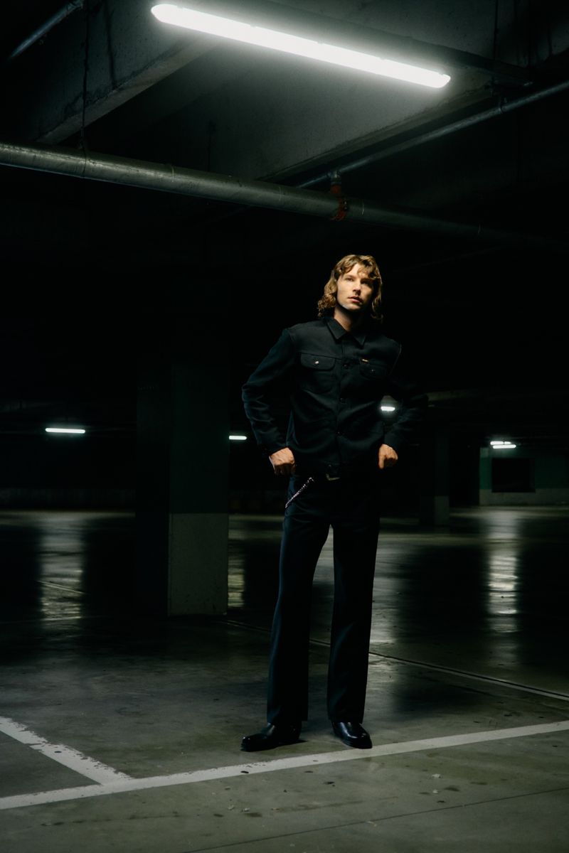 A man with long hair wearing a dark denim jacket and matching trousers stands in a dimly lit underground parking garage.