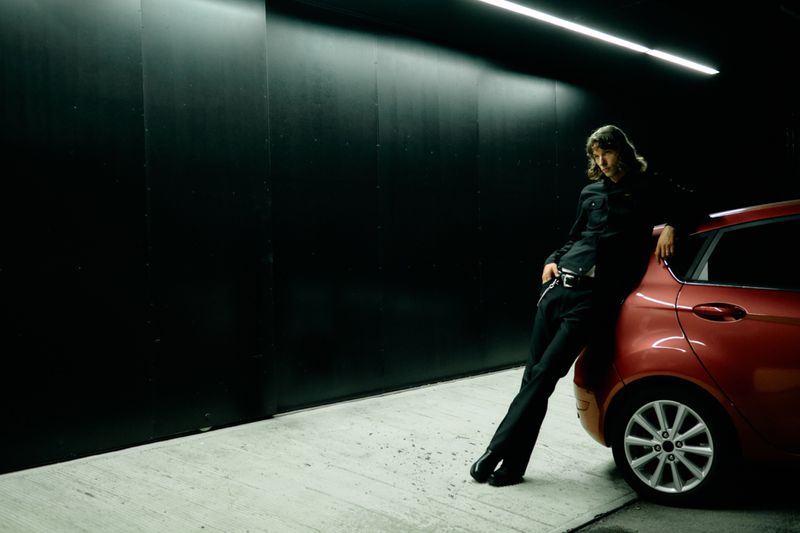 Model leaning against a red car in a dark parking garage under a linear overhead light.