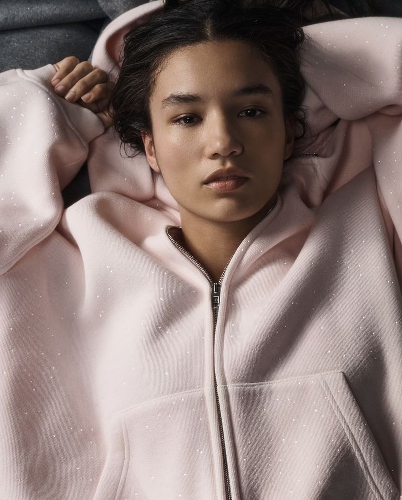 Close-up portrait of a model lying down wearing a pale pink zip-up hoodie with subtle speckles