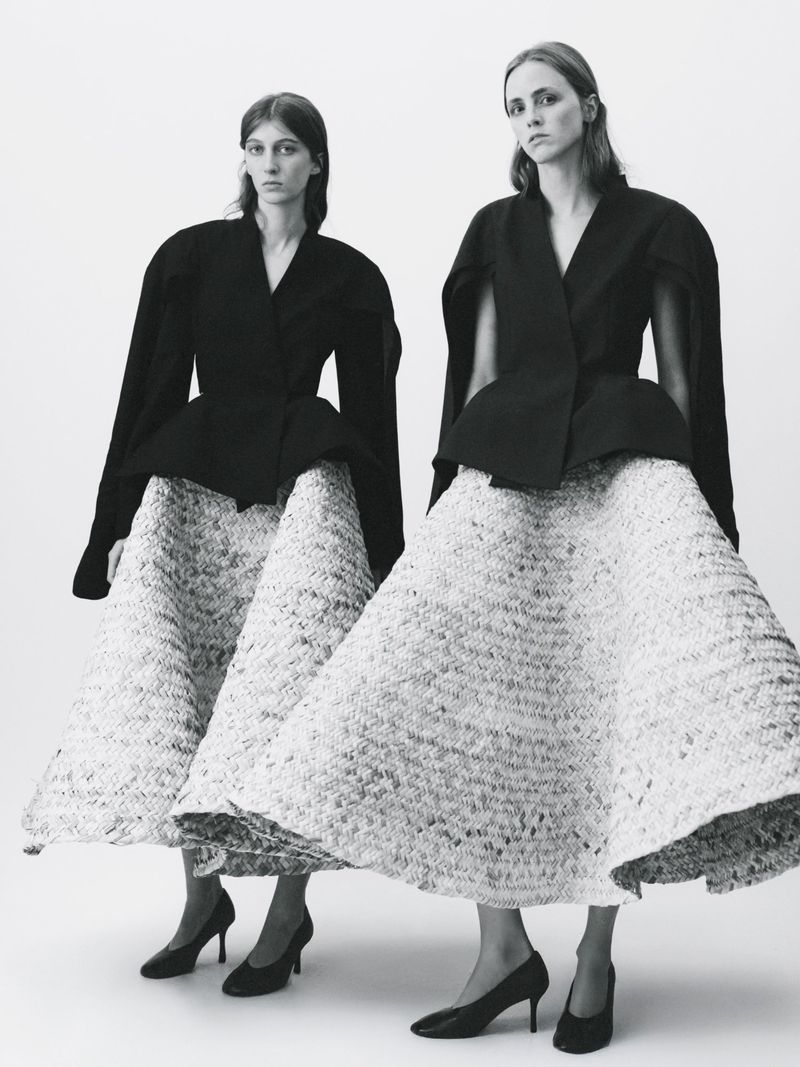 Two female models wearing structured black jackets and voluminous textured skirts standing against a plain white background