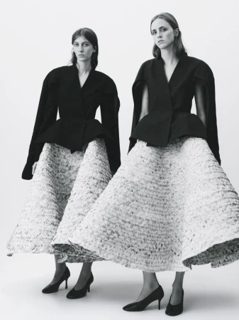 Two female models wearing structured black jackets and voluminous textured skirts standing against a plain white background