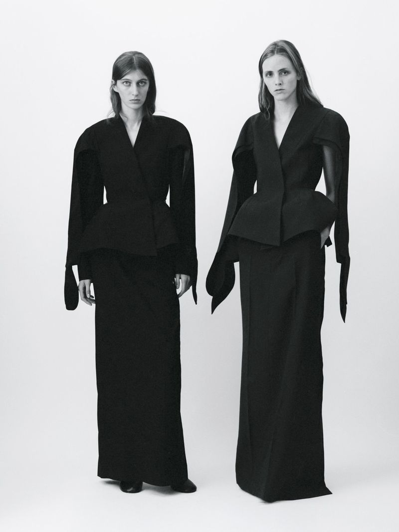 Two female models wearing black structured jackets and long skirts against a plain white studio background.