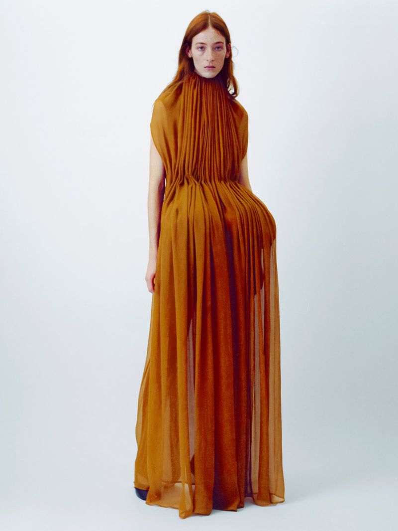 Model with long red hair wearing a pleated amber gown against a plain white studio background.