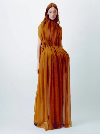 Model with long red hair wearing a pleated amber gown against a plain white studio background.