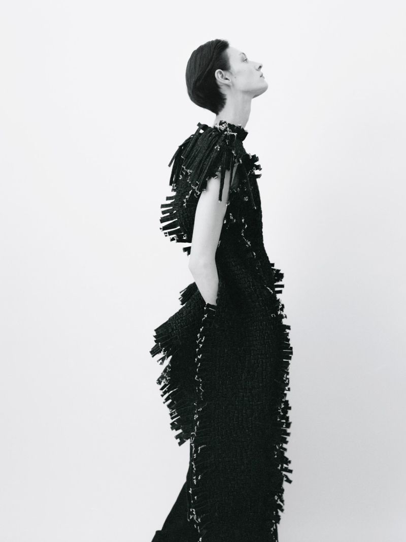 Model in profile wearing a black textured sleeveless dress with fringe details against a white background