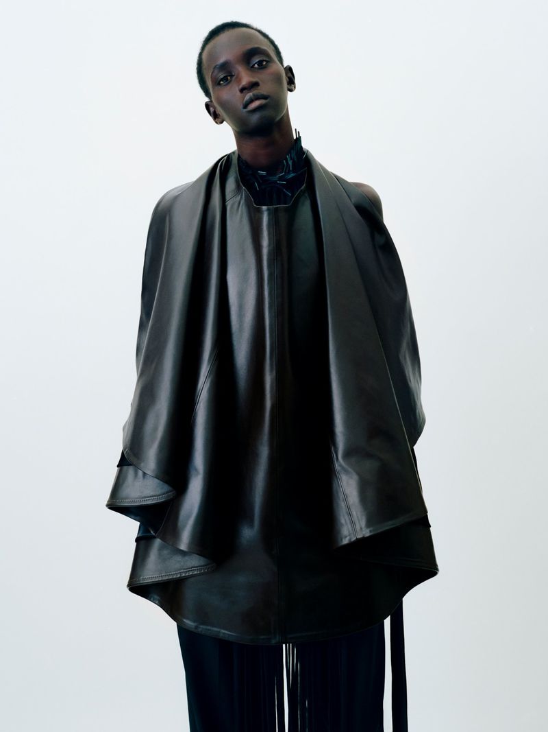 Model wearing a structured dark leather cape and top against a plain white background for Institution By Galib Gassanoff