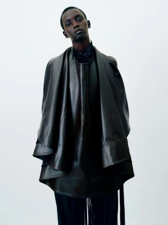 Model wearing a structured dark leather cape and top against a plain white background for Institution By Galib Gassanoff