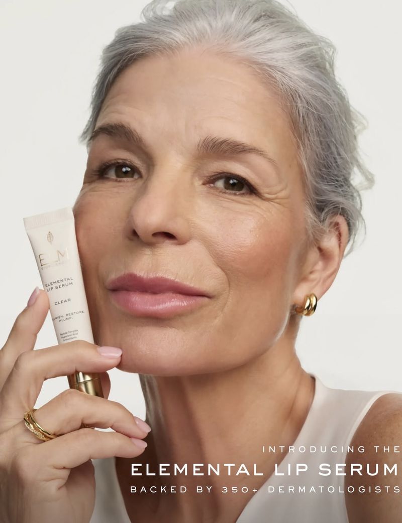 Close-up portrait of an elderly woman holding a lip serum tube against a plain white background