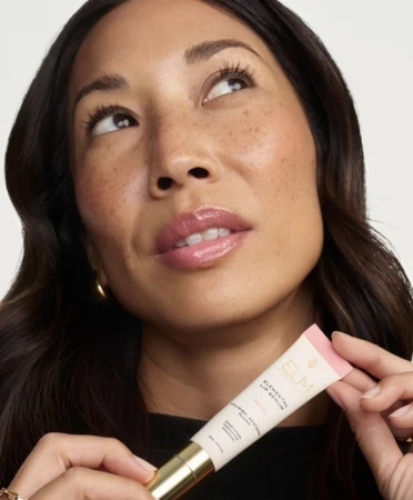 Close-up portrait of a woman holding a tube of Elm Biosciences Elemental Lip Serum against a white background