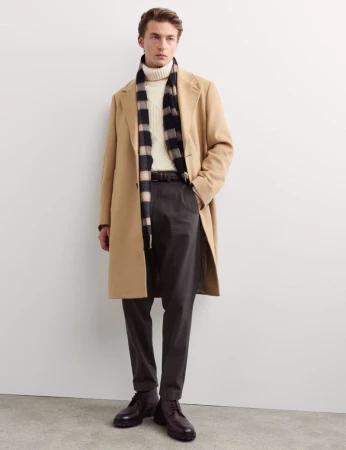 Model wearing a camel overcoat, cream turtleneck sweater, checkered scarf, and dark trousers against a white seamless background