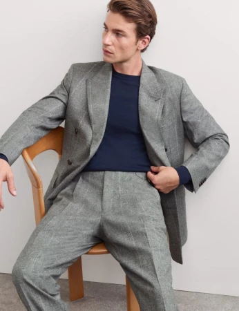 Man wearing a grey tailored double-breasted suit jacket and matching trousers seated on a wooden chair against a plain white background
