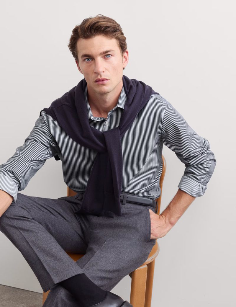 Male model seated on a wooden chair wearing a striped shirt, grey trousers, and a navy sweater draped over his shoulders