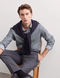 Male model seated on a wooden chair wearing a striped shirt, grey trousers, and a navy sweater draped over his shoulders