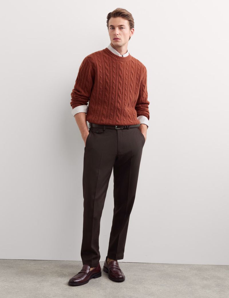 Man wearing a rust cable knit sweater and dark trousers posing against a clean white studio background