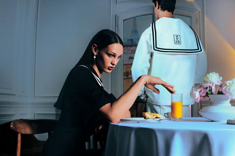 Stylish editorial shot featuring a young woman in a dark dress with a sailor collar seated at a table with a man in a sailor suit.