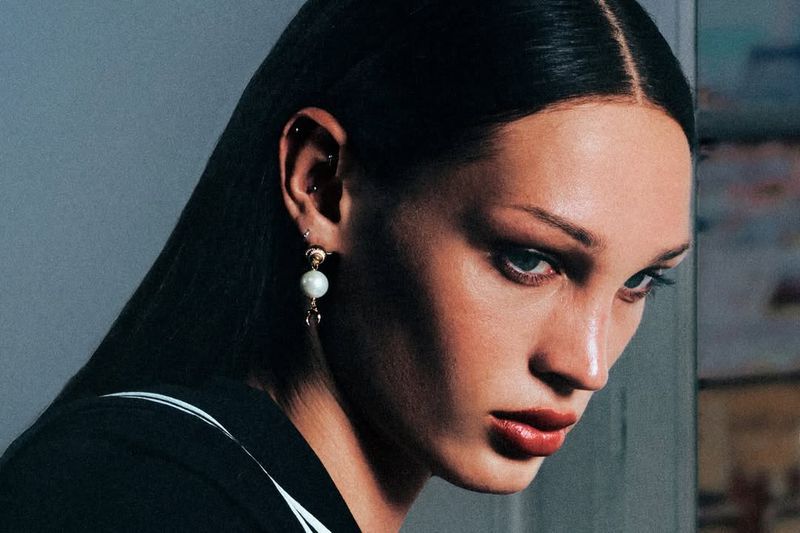 Portrait of a woman with dark hair wearing a black blazer with a white stripe, featuring bright red lipstick and pearl earrings.