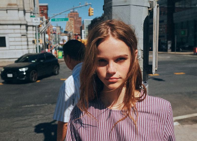 Model with long brown hair wearing a striped top standing outdoors on a city street next to a pole.