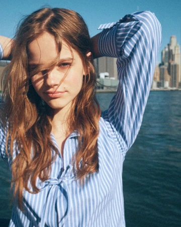 Young woman with long brown hair wearing a blue and white striped shirt outdoors by water with city buildings in the background.