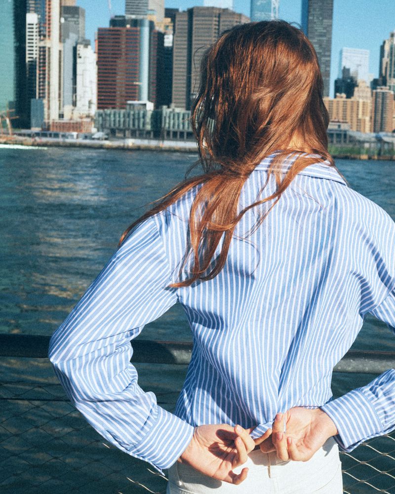 Model with long reddish-brown hair wearing a blue and white striped shirt outdoors by a waterfront