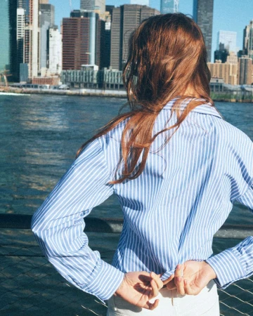 Model with long reddish-brown hair wearing a blue and white striped shirt outdoors by a waterfront