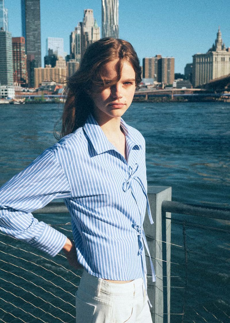 Model wearing a blue and white striped shirt with bow details standing outdoors against an urban waterfront skyline.