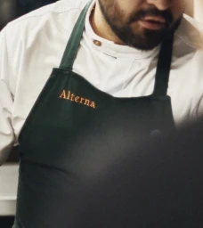 Chef wearing a white chef coat and a dark green apron with embroidered orange Alterna branding.