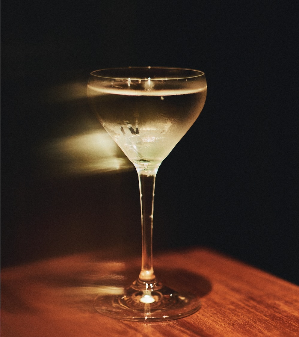 A chilled cocktail in a stemmed coupe glass resting on a wooden surface against a dark background with light streaks.