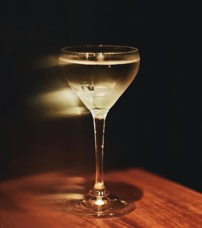 A chilled cocktail in a stemmed coupe glass resting on a wooden surface against a dark background with light streaks.