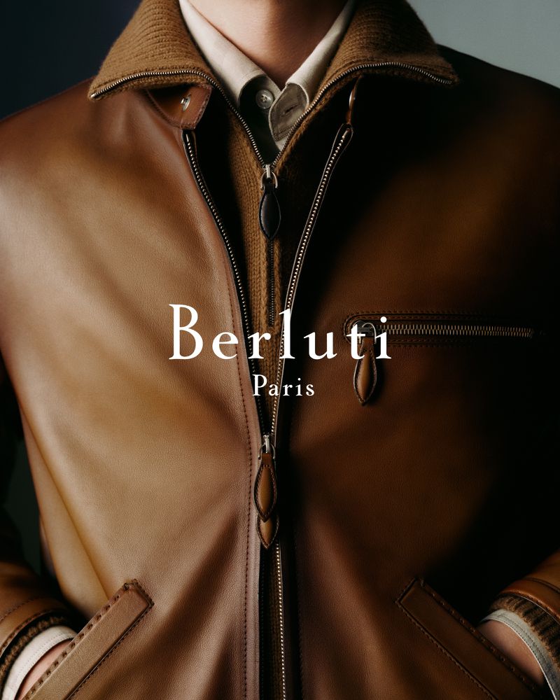 Close-up of a man wearing a brown leather jacket with zipper. Berluti brand logo visible on the left. Fashion advertisement.