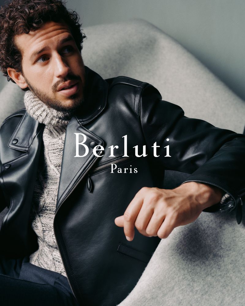A curly-haired man in a black leather jacket poses indoors with the 'Berluti Paris' logo, showcasing fashion apparel against a light gray backdrop.