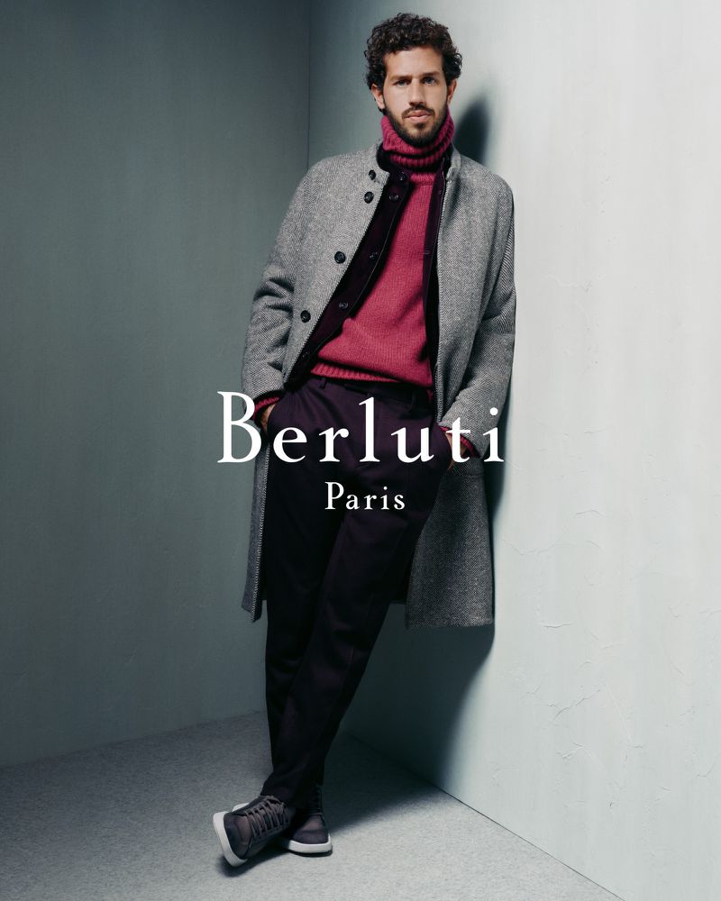 A man in a grey overcoat, maroon turtleneck sweater, and dark pants leans against a wall in a studio, showcasing a modern, upscale look.