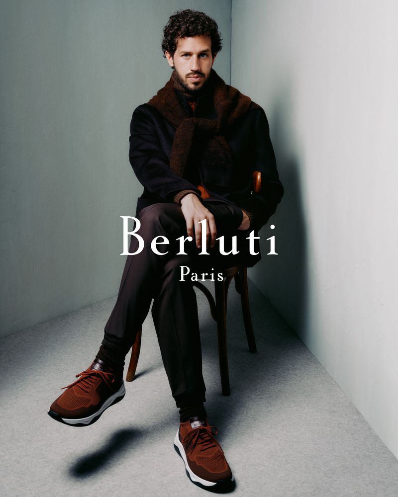 Man in a coat and brown athletic shoes sits on a wooden chair in a studio. The text overlay displays the Berluti Paris brand.