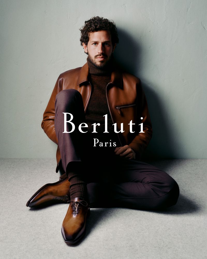 Portrait of a handsome man in a brown leather jacket and brown pants sitting on the floor, with Berluti Paris logo.