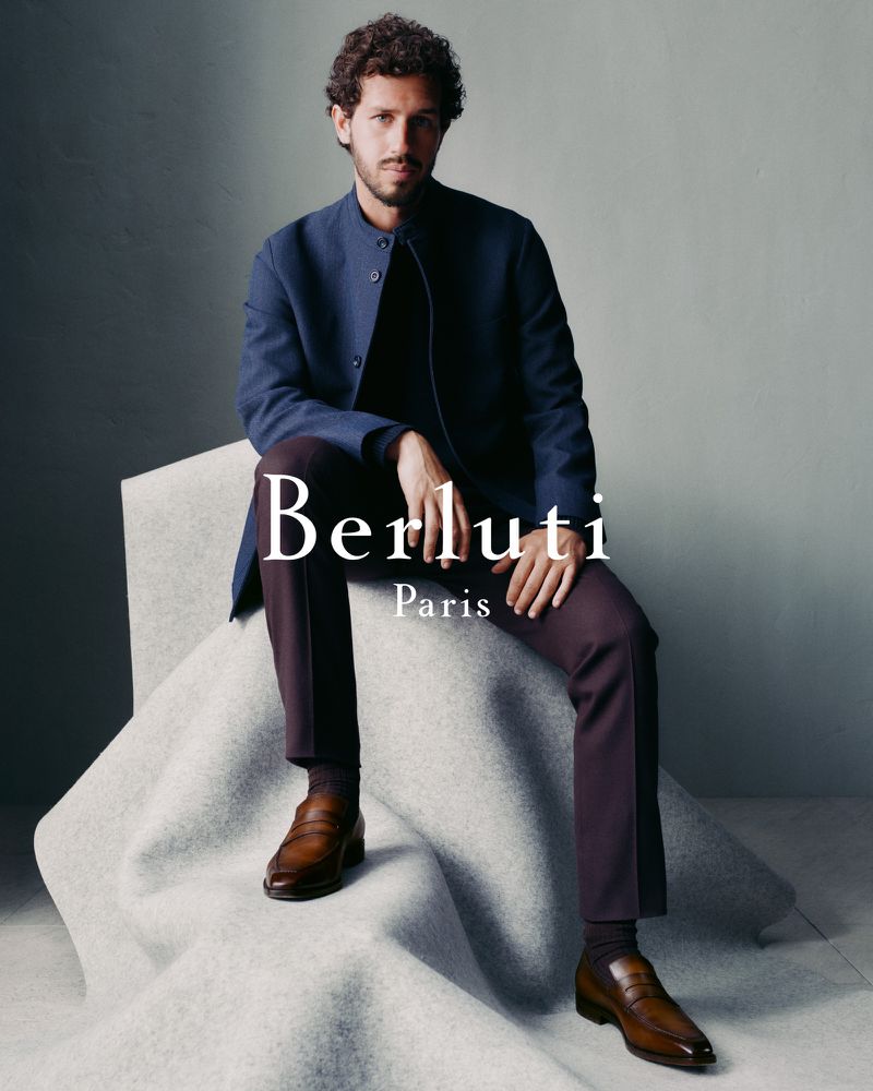 A man in a blue jacket and brown trousers is seated on a grey draped cube, wearing brown leather shoes. The backdrop is a plain grey.