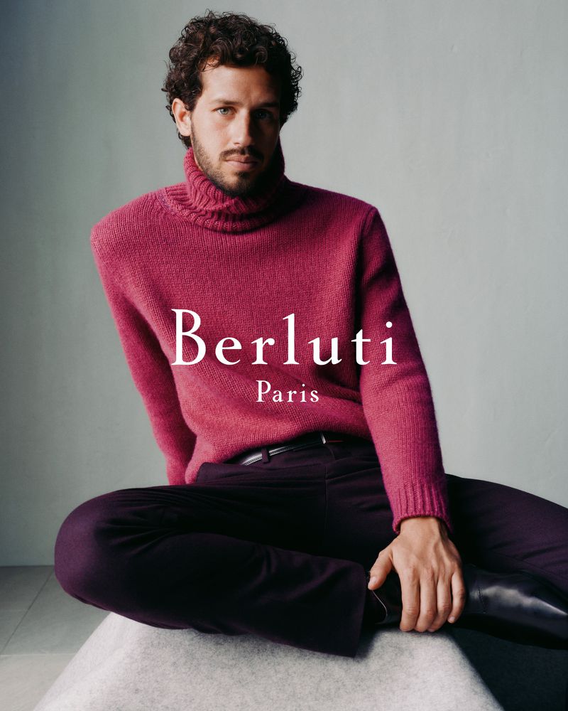 Man in a magenta turtleneck sweater and dark trousers poses sitting on a stool in a studio for Berluti Paris fashion ad.